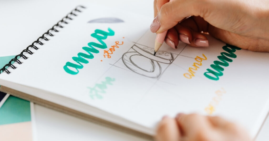 A person sketching hand-lettered typography in a notebook, surrounded by brand colour swatches – representing the creative process of choosing the right font for brand identity.